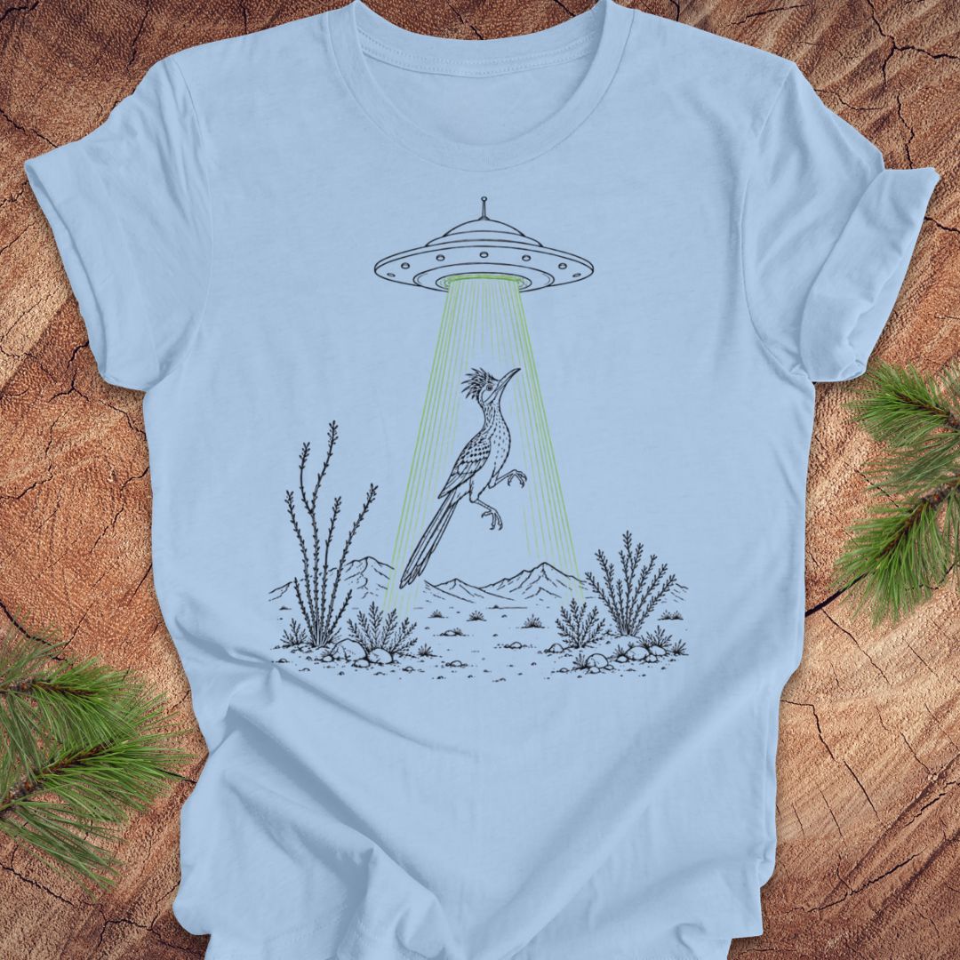 A light blue crewneck t-shirt laid flat on a rustic wood background with pine branches in the corners. The t-shirt features a graphic illustration of a gray bird under a UFO with a green beam of light shining down.