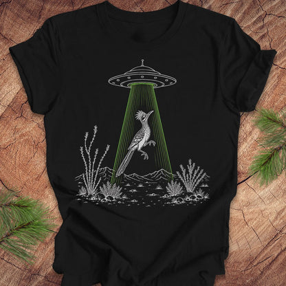 A black short-sleeve t-shirt laid flat on a rustic wood surface, featuring a white line-art graphic of a bird under a UFO with a bright green beam of light. Green pine branches are placed around the shirt as styling props.