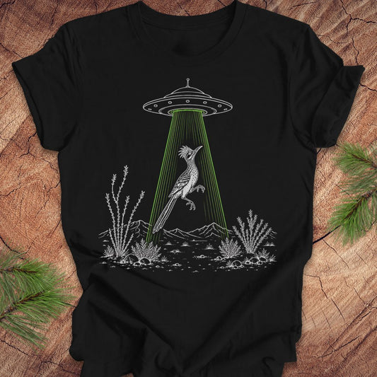 A black short-sleeve t-shirt laid flat on a rustic wood surface, featuring a white line-art graphic of a bird under a UFO with a bright green beam of light. Green pine branches are placed around the shirt as styling props.