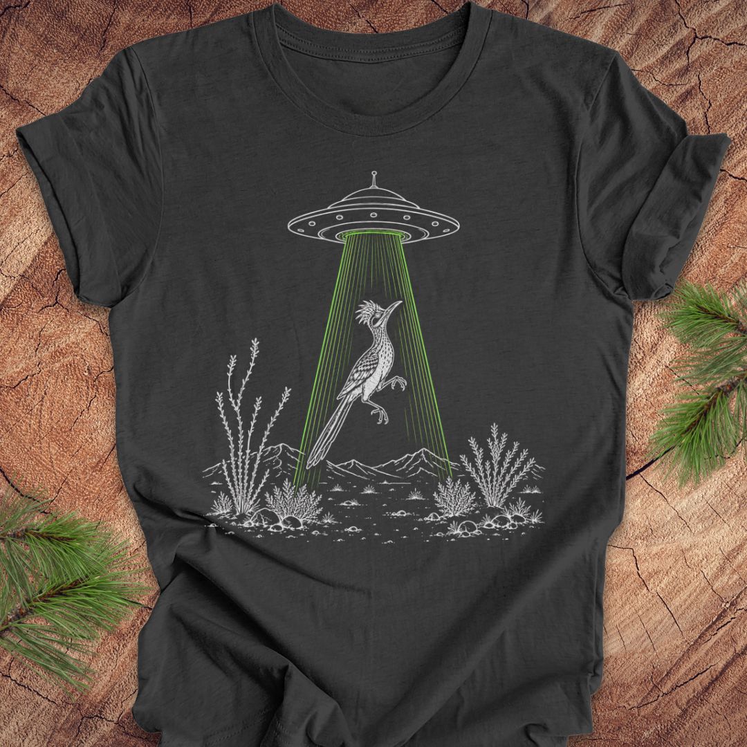 A dark charcoal gray t-shirt with a graphic of a white bird under a green laser beam from a UFO, laid flat on a rustic wood background with pine branches.