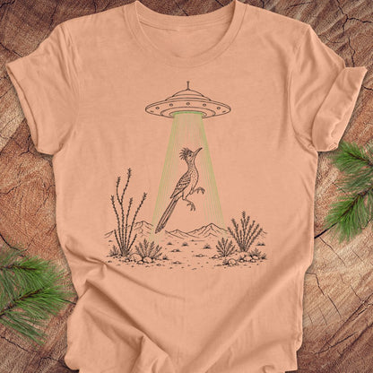 A peach-colored short-sleeve t-shirt laid flat on a rustic wood background with pine branches. The shirt features a graphic of a desert bird under a UFO with green beam rays shining down.