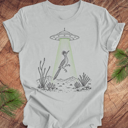 A light gray short-sleeve t-shirt laid flat on a rustic wood surface, featuring a black line art graphic of a bird under a UFO with green rays shining down. Pine branches are visible in the corners of the frame.