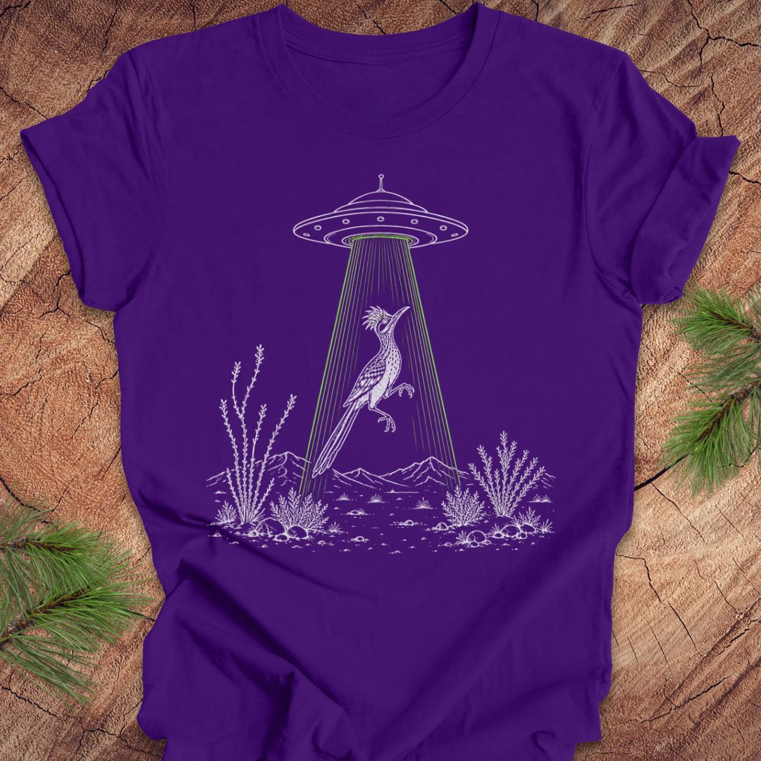 A purple short-sleeve crewneck t-shirt laid flat on a rustic wood background with pine branches in the corners. The shirt features a white line-art graphic of a bird under a UFO with green light rays shining down.