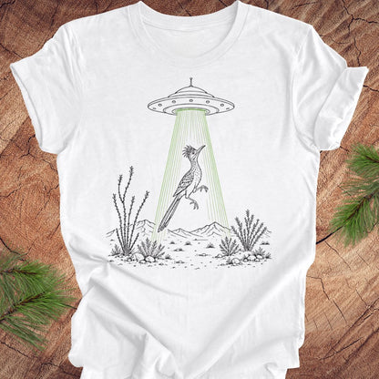 A white short-sleeve crewneck t-shirt laid flat on a rustic wood background with pine branches in the corners. The t-shirt features a black line art graphic of a bird under a flying saucer with green rays shining down.