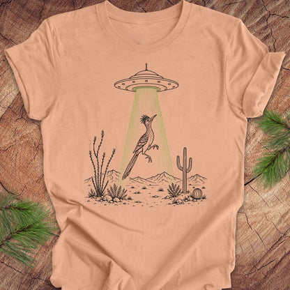 A peach-colored short-sleeve t-shirt laid flat on a rustic wood background, featuring a graphic of a desert bird under a UFO with green light rays.