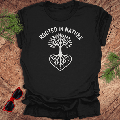 Black crew neck t-shirt with a white graphic of a tree with roots and the phrase 'ROOTED IN NATURE' printed above it, displayed on a wooden surface with pine needles and red sunglasses to the side.