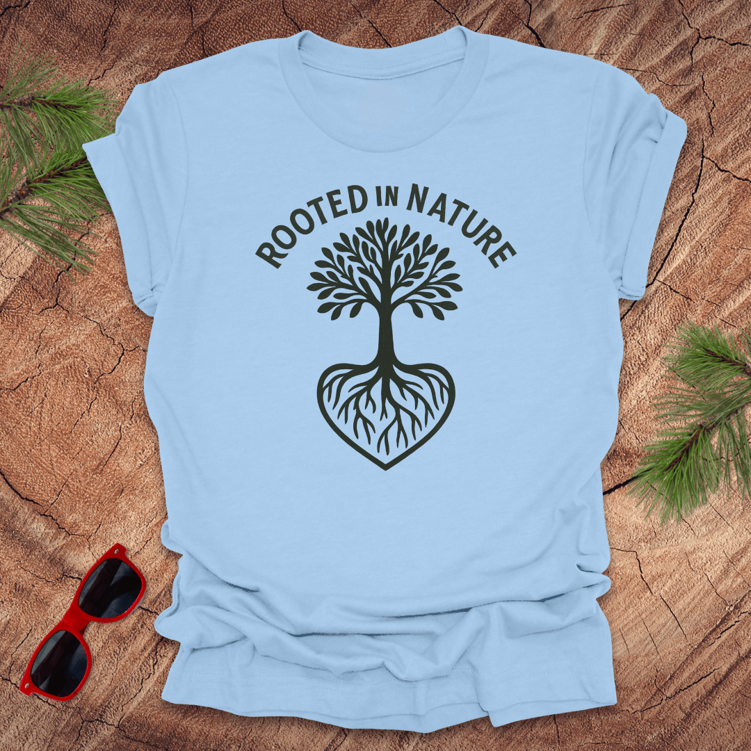 Blue crew neck t-shirt with a white graphic of a tree with roots and the phrase 'ROOTED IN NATURE' printed above it, displayed on a wooden surface with pine needles and red sunglasses to the side.