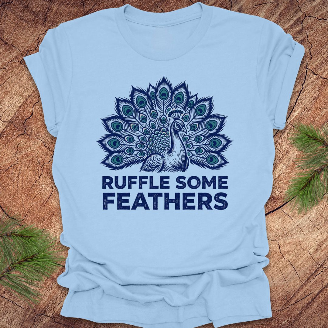 Light blue t-shirt with peacock design and 'Ruffle Some Feathers' text on a wooden surface.