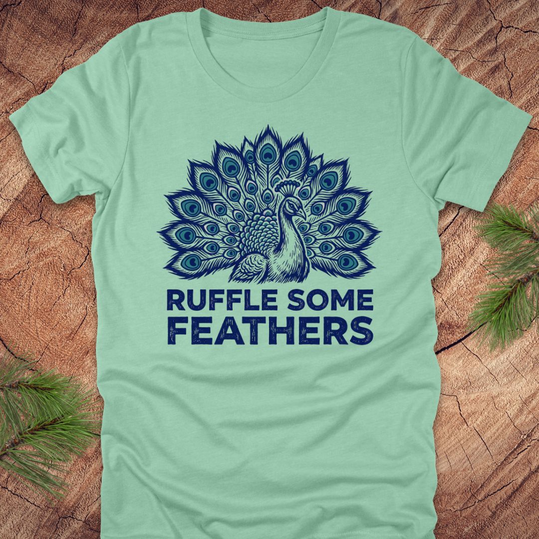 Light green t-shirt with a peacock design and 'Ruffle Some Feathers' text on a wooden surface.