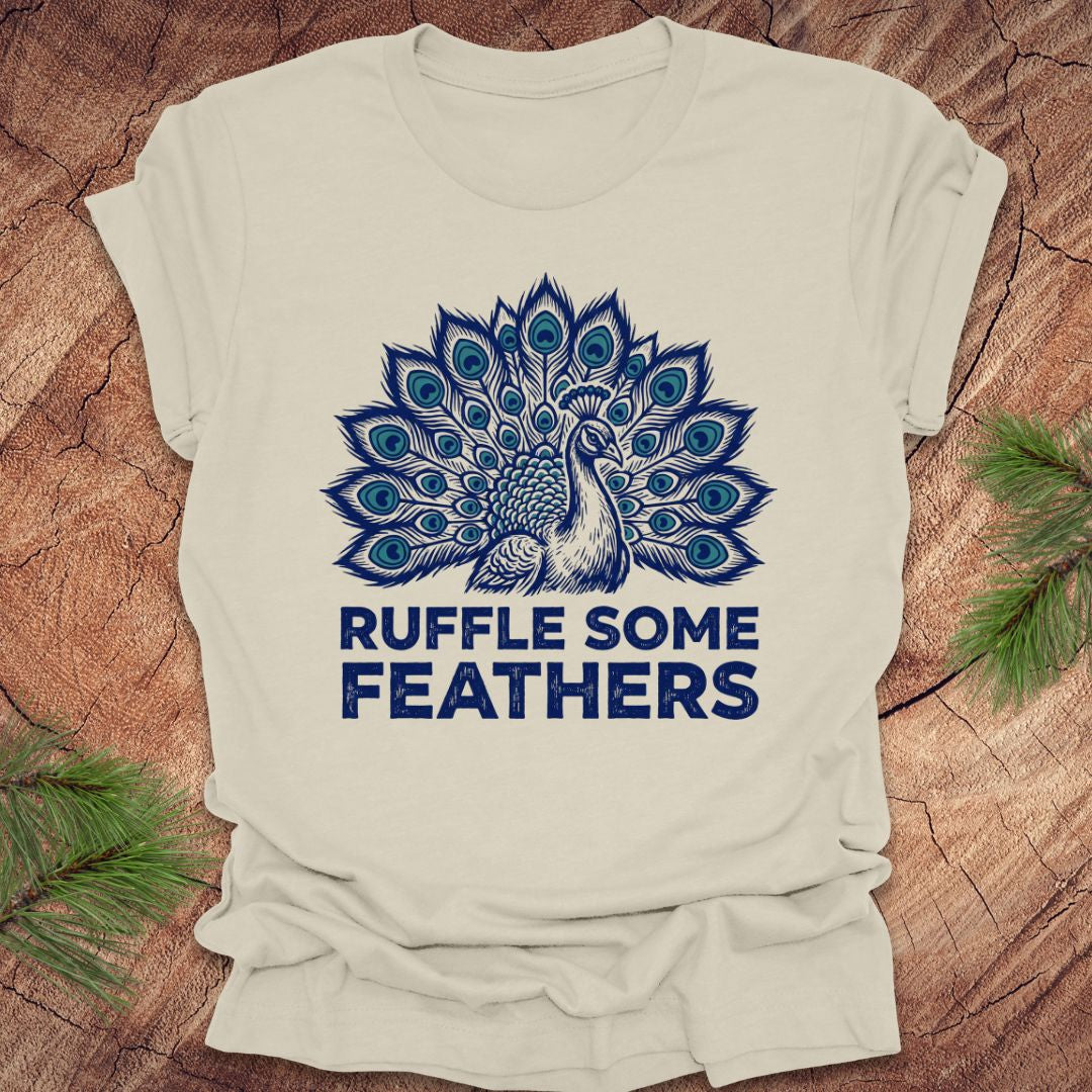Beige t-shirt with blue peacock design and 'Ruffle Some Feathers' text on a wooden surface.