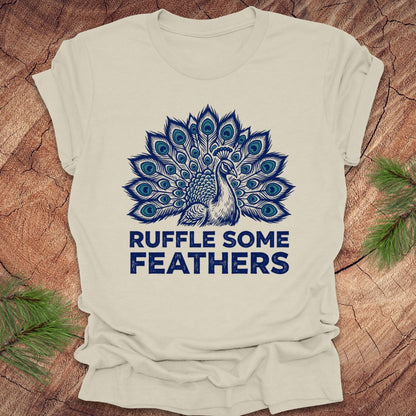 Beige t-shirt with blue peacock design and 'Ruffle Some Feathers' text on a wooden surface.