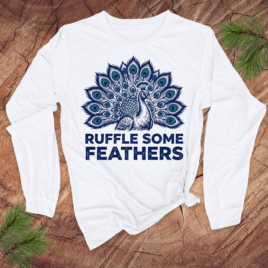 White long-sleeve shirt with peacock design and 'Ruffle Some Feathers' text on a wooden surface.