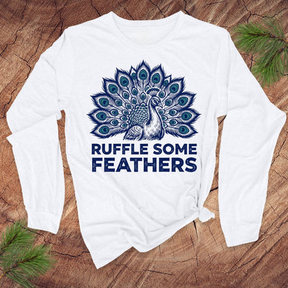 White long-sleeve shirt with peacock design and 'Ruffle Some Feathers' text on a wooden surface.