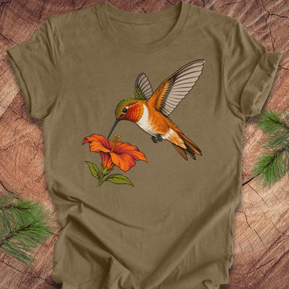 Heather olive t-shirt with a Rufous Hummingbird hovering at an orange trumpet flower, laid flat on a rustic wood background with pine branches.
