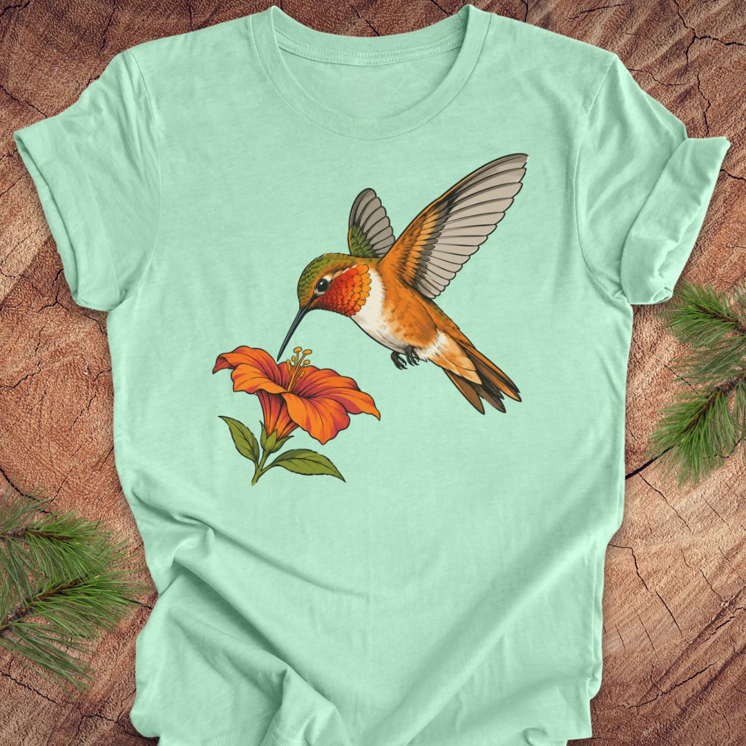 Heather mint green t-shirt with a Rufous Hummingbird hovering at an orange trumpet flower, laid flat on a rustic wood background with pine branches.