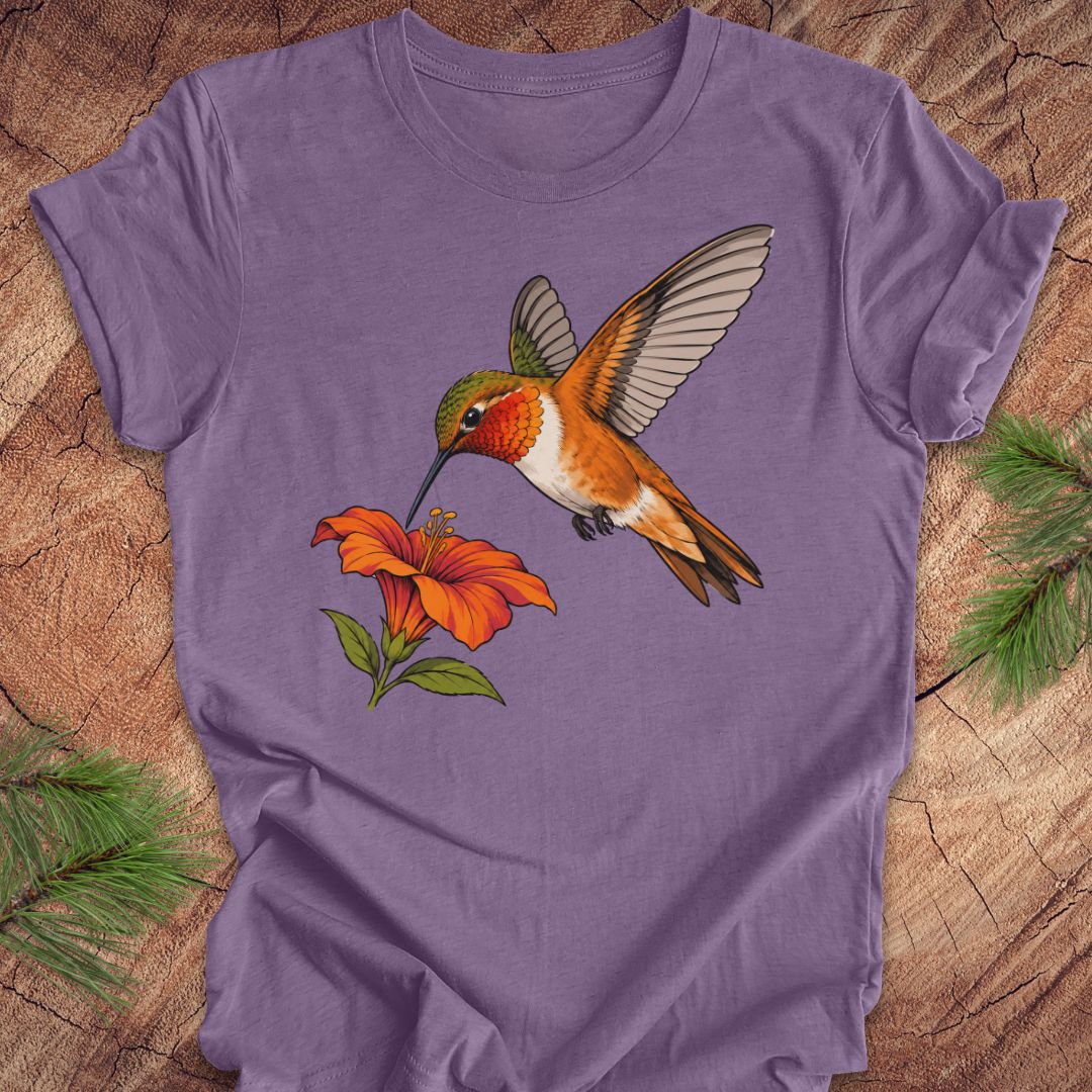 Heather purple t-shirt with a Rufous Hummingbird hovering at an orange trumpet flower, laid flat on a rustic wood background with pine branches.