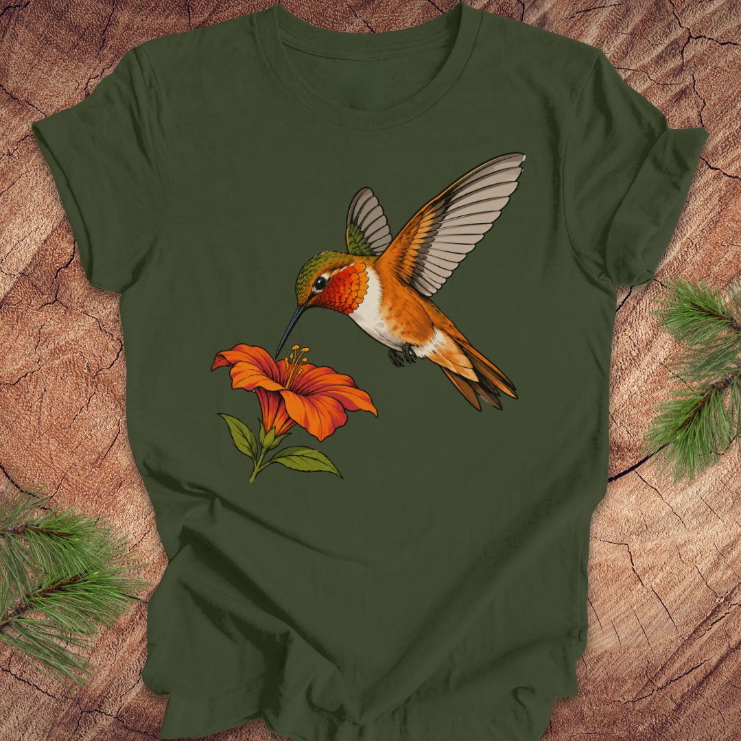 Military green t-shirt with a Rufous Hummingbird hovering at an orange trumpet flower, laid flat on a rustic wood background with pine branches.