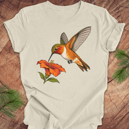 Natural cream t-shirt with a Rufous Hummingbird hovering at an orange trumpet flower, laid flat on a rustic wood background with pine branches.
