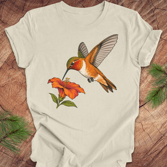 Natural cream t-shirt with a Rufous Hummingbird hovering at an orange trumpet flower, laid flat on a rustic wood background with pine branches.