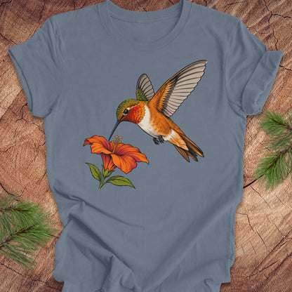 Steel blue t-shirt with a Rufous Hummingbird hovering at an orange trumpet flower, laid flat on a rustic wood background with pine branches.