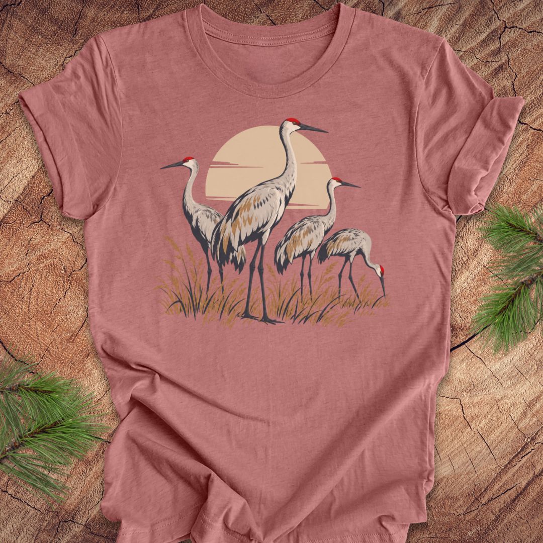 A dusty rose short-sleeve t-shirt featuring a graphic of three cranes in front of a warm sun, laid flat on a rustic wood background with pine needles in the corners.