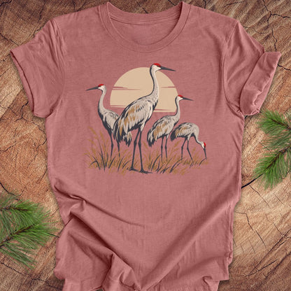 A dusty rose short-sleeve t-shirt featuring a graphic of three cranes in front of a warm sun, laid flat on a rustic wood background with pine needles in the corners.
