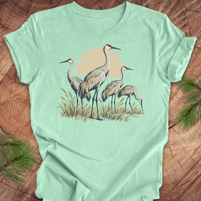 A mint green short-sleeve t-shirt laid flat on a rustic wood background, featuring a graphic print of a family of sandhill cranes standing in reeds against a pale sun.