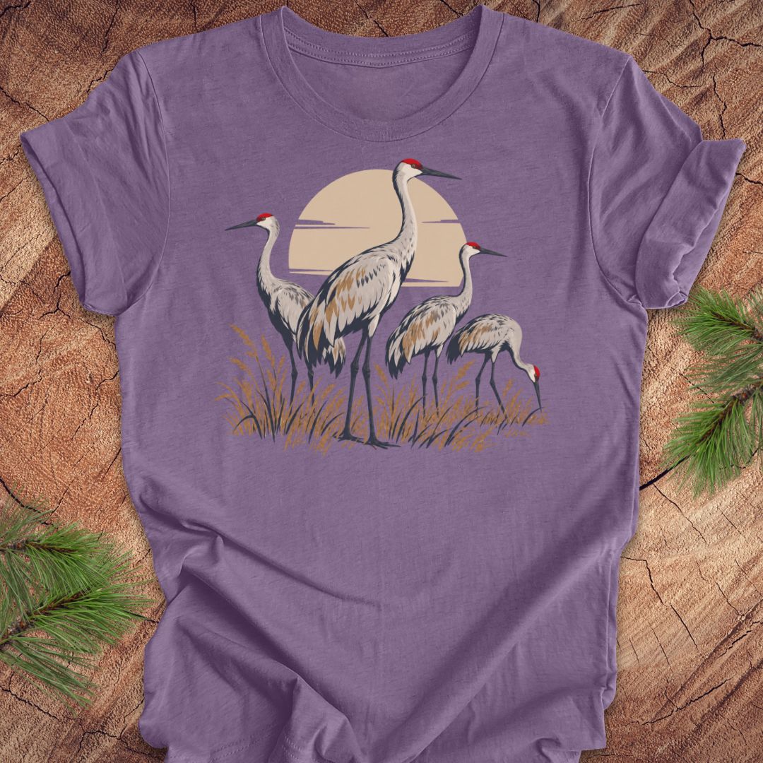 A purple short-sleeve crewneck t-shirt featuring a graphic of three sandhill cranes in front of a beige sun, laid flat on a rustic wood background with pine needles in the corners.