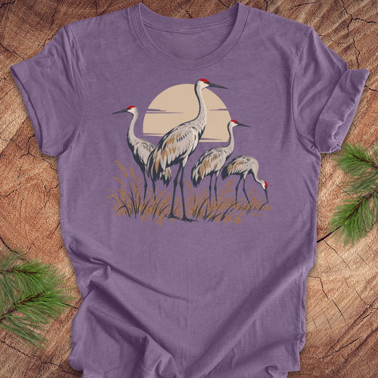 A purple short-sleeve crewneck t-shirt featuring a graphic of three sandhill cranes in front of a beige sun, laid flat on a rustic wood background with pine needles in the corners.