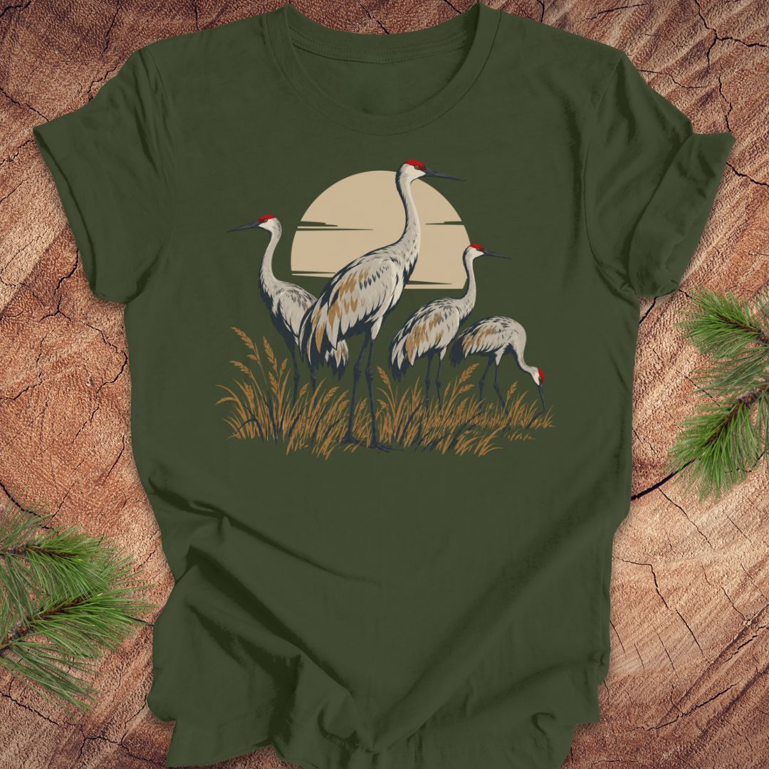 An olive green short-sleeve t-shirt featuring a graphic of three sandhill cranes in front of a beige sun over grass, laid flat on a rustic wood background with pine needles in the corners.