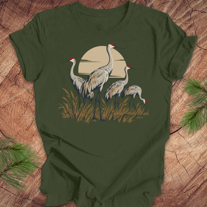 An olive green short-sleeve t-shirt featuring a graphic of three sandhill cranes in front of a beige sun over grass, laid flat on a rustic wood background with pine needles in the corners.