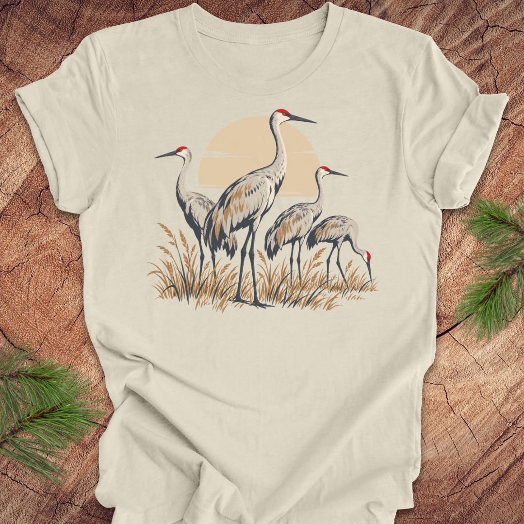 An off-white short-sleeve t-shirt featuring a graphic print of three sand cranes standing in reeds against a pale orange sun, laid flat on a rustic wood background with pine needles in the corners.