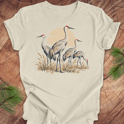 An off-white short-sleeve t-shirt featuring a graphic print of three sand cranes standing in reeds against a pale orange sun, laid flat on a rustic wood background with pine needles in the corners.