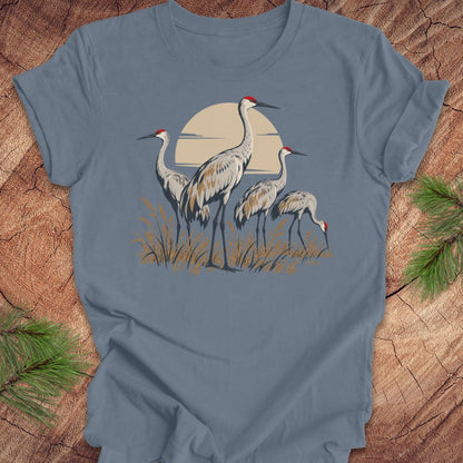 A slate blue short-sleeve t-shirt with a graphic of three sandhill cranes in front of a large beige sun, laid flat on a rustic wood background with pine needles in the corners.