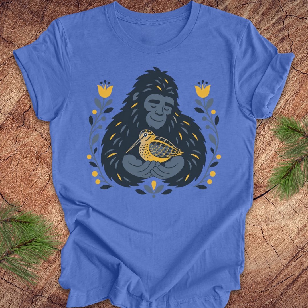 A blue short-sleeve crewneck t-shirt laid flat on a rustic wood background, featuring a graphic of a sasquatch holding a woodcock bird, surrounded by yellow flowers and green leaves.