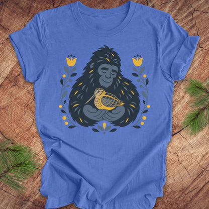 A blue short-sleeve crewneck t-shirt laid flat on a rustic wood background, featuring a graphic of a sasquatch holding a woodcock bird, surrounded by yellow flowers and green leaves.