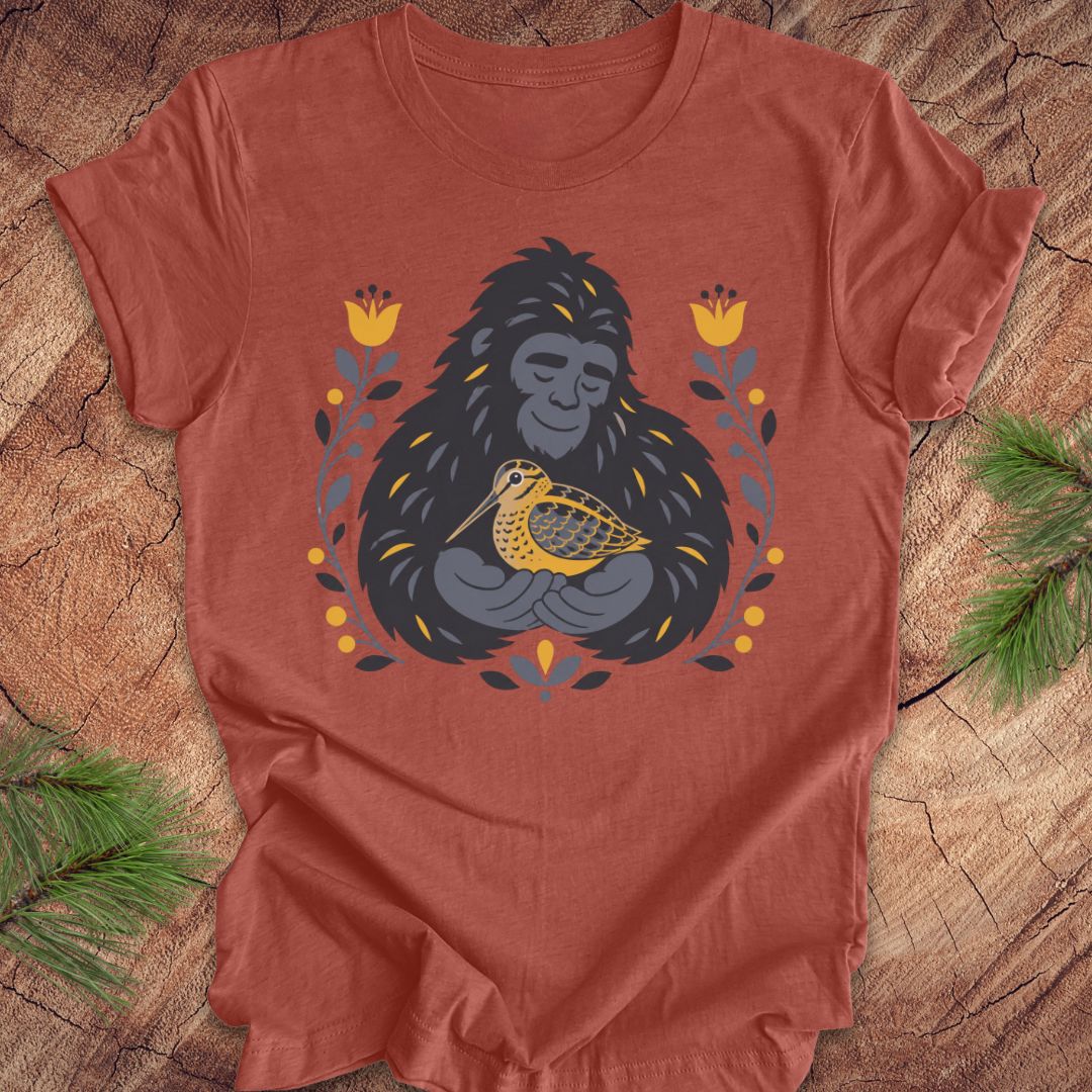 A terracotta colored short-sleeve crewneck t-shirt laid flat on a rustic wood background, featuring a graphic of a sasquatch holding a woodcock bird, surrounded by yellow flowers and green leaves.