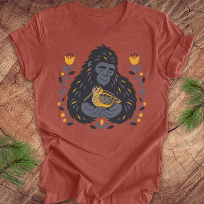 A terracotta colored short-sleeve crewneck t-shirt laid flat on a rustic wood background, featuring a graphic of a sasquatch holding a woodcock bird, surrounded by yellow flowers and green leaves.