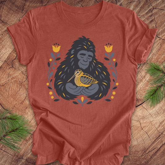 A terracotta colored short-sleeve crewneck t-shirt laid flat on a rustic wood background, featuring a graphic of a sasquatch holding a woodcock bird, surrounded by yellow flowers and green leaves.