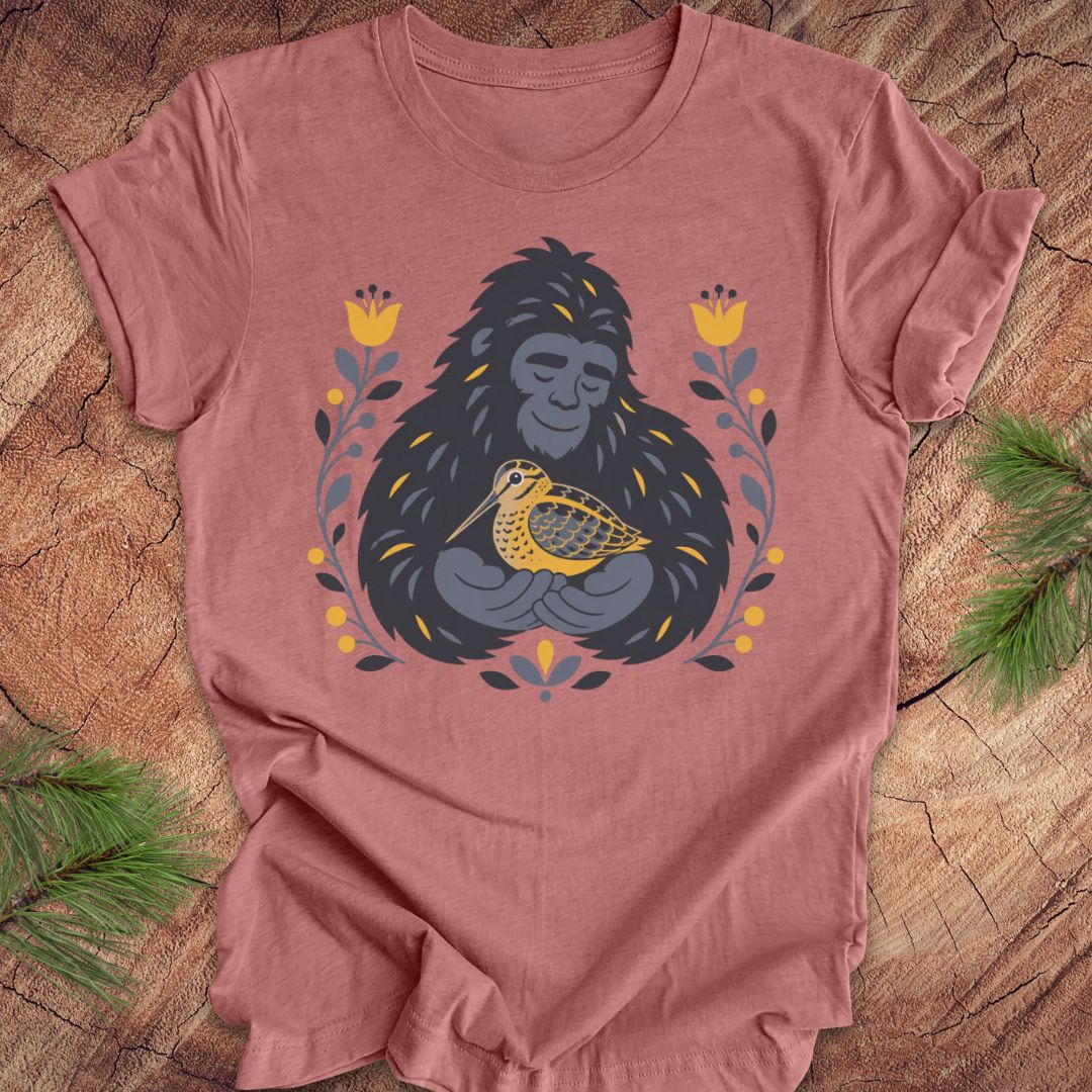 A mauve short-sleeve crewneck t-shirt laid flat on a rustic wood background, featuring a graphic of a sasquatch holding a woodcock bird, surrounded by yellow flowers and green leaves.