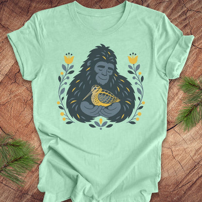 A mint green short-sleeve crewneck t-shirt laid flat on a rustic wood background, featuring a graphic of a sasquatch holding a woodcock bird, surrounded by yellow flowers and green leaves.