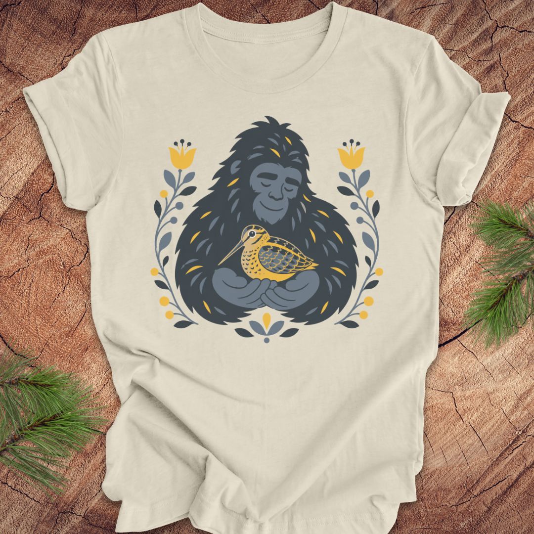 A beige short-sleeve crewneck t-shirt laid flat on a rustic wood background, featuring a graphic of a sasquatch holding a woodcock bird, surrounded by yellow flowers and green leaves.