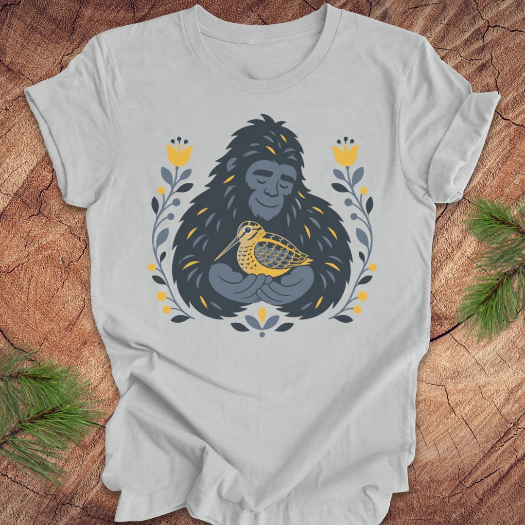 A light gray short-sleeve crewneck t-shirt laid flat on a rustic wood background, featuring a graphic of a sasquatch holding a woodcock bird, surrounded by yellow flowers and green leaves.