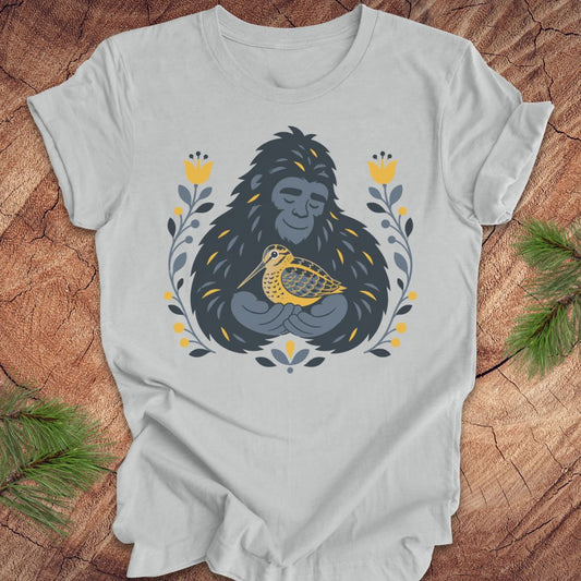 A light gray short-sleeve crewneck t-shirt laid flat on a rustic wood background, featuring a graphic of a sasquatch holding a woodcock bird, surrounded by yellow flowers and green leaves.