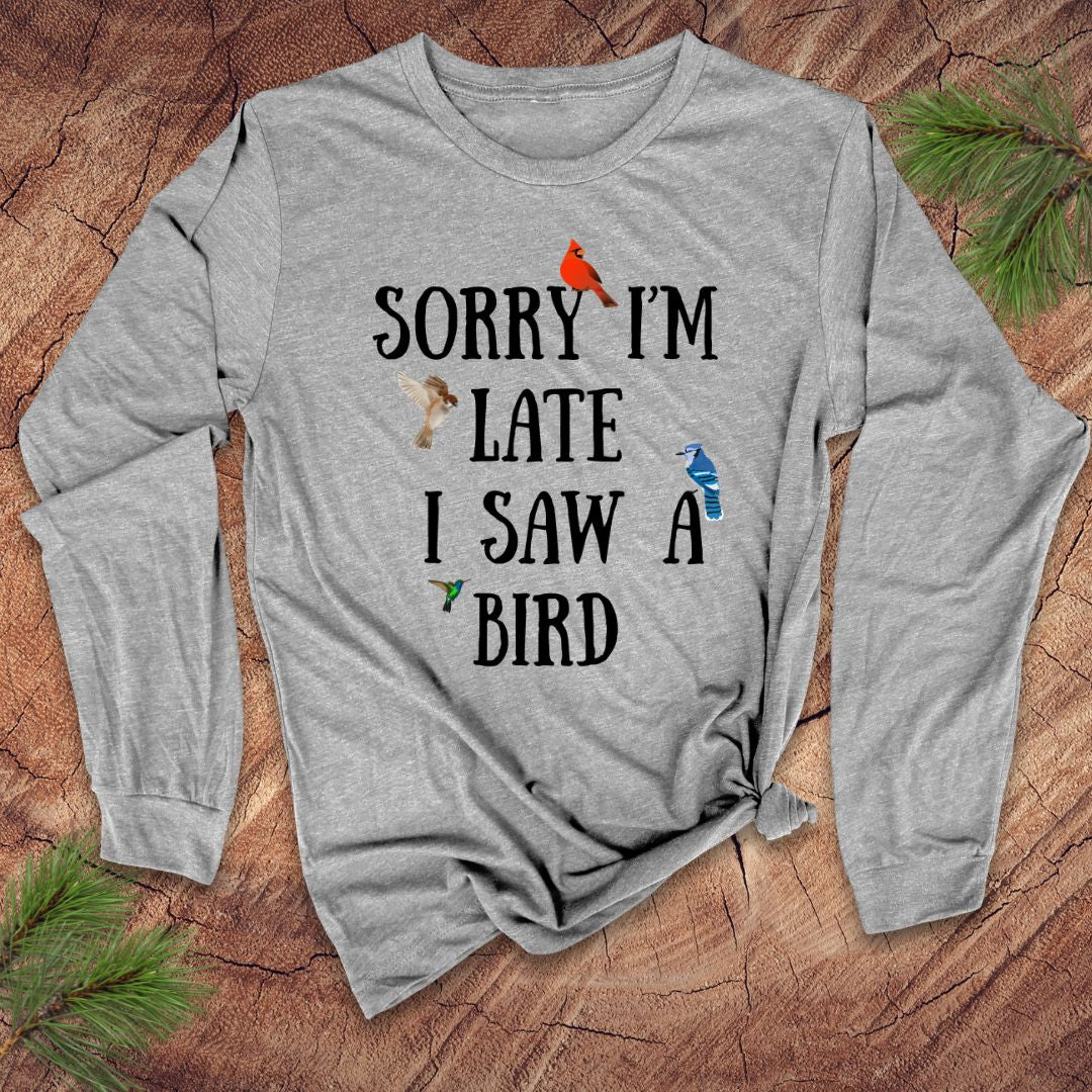 Light gray long-sleeve shirt with text 'Sorry I'm late I saw a bird' on a wooden surface with pine branches.