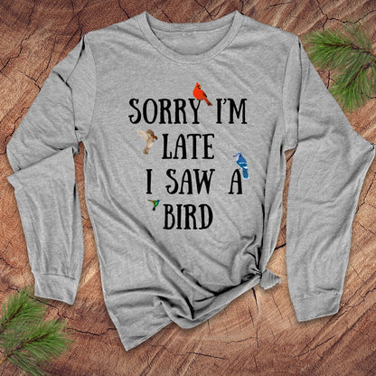 Light gray long-sleeve shirt with text 'Sorry I'm late I saw a bird' on a wooden surface with pine branches.