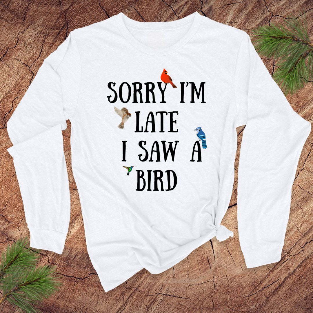 White long sleeve tshirt with text 'Sorry I'm late I saw a bird' on a wooden surface with greenery.