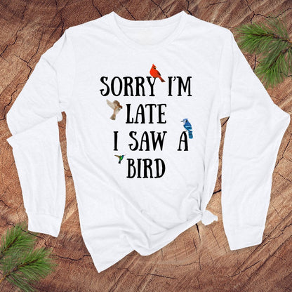 White long sleeve tshirt with text 'Sorry I'm late I saw a bird' on a wooden surface with greenery.