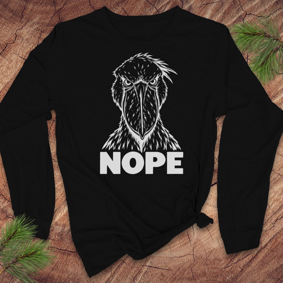Black long-sleeve t-shirt with a white line-art Shoebill staring straight ahead above the word NOPE, laid flat on a rustic wood background with pine branches.