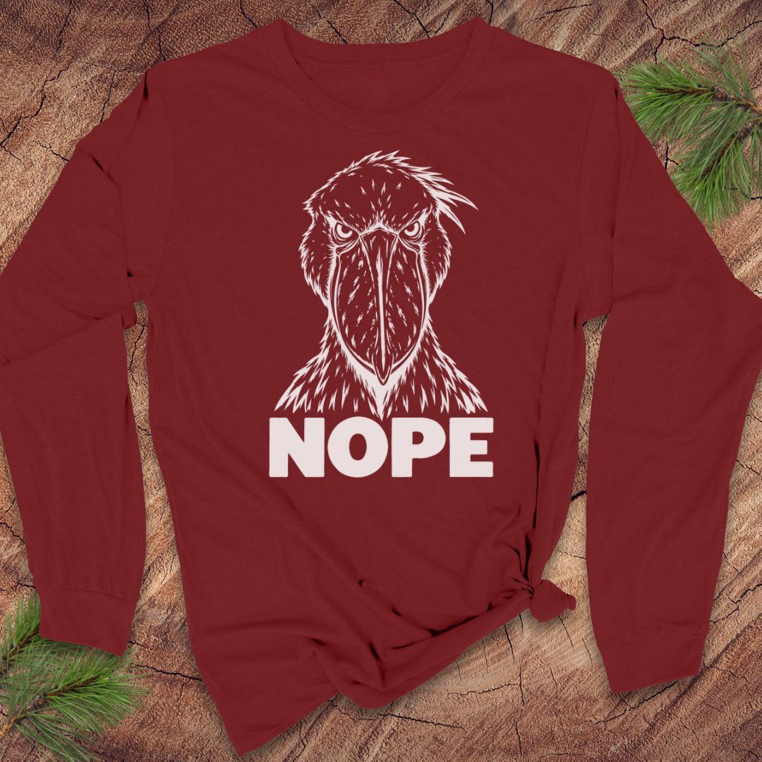 Cardinal red long-sleeve t-shirt with a white line-art Shoebill staring straight ahead above the word NOPE, laid flat on a rustic wood background with pine branches.